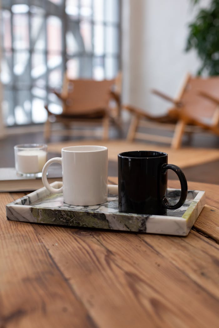 Two ceramic mugs, black and white, on a marble tray indoors, with a cozy background.
